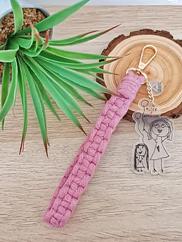 Keychain with a pink macramé strap and a clear acrylic charm printed with a child's drawing, cut around its outline.