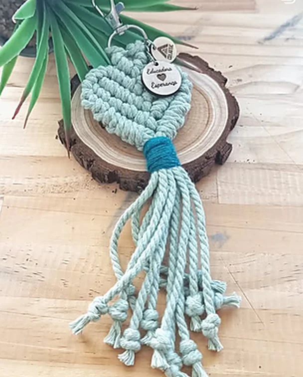 Mint macramé heart keychain with a fringe and an engraved wooden tag, on a log slice.