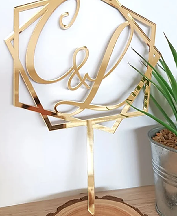 Gold mirror acrylic cake topper with two calligraphic initials inside a geometric frame.